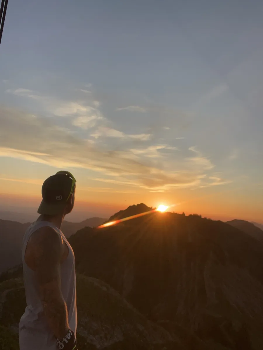 Thomas von hinten mit Kappe, Blick auf den Sonnenuntergang über einer Bergkette